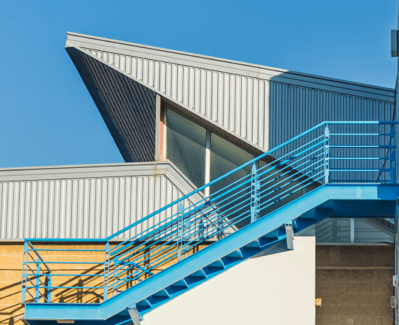 Modern building exterior with a bright blue metal staircase, a gray angular roof, and corrugated metal siding.