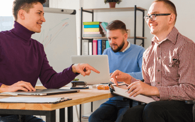 Three men working together in an office, one using a wheelchair, collaborating around a table with a laptop and documents.
