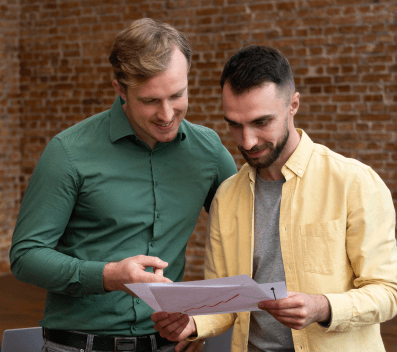 Two men looking at a document with a graph on it, standing in front of a brick wall.